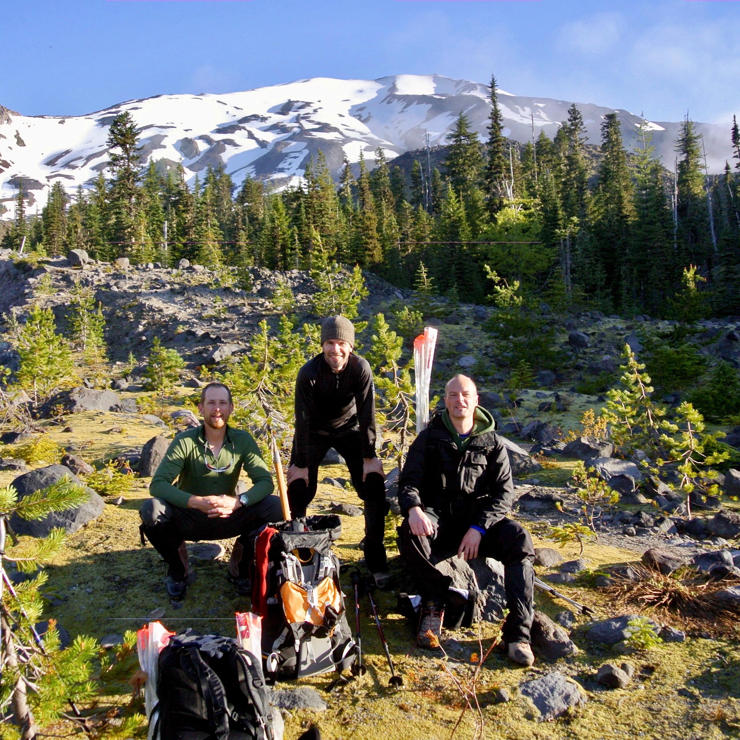 Matthew Kessi (right) and two friends prepare to climb to the summit of Mt. St. Helens in Washington State.  Two of them are seated while the third hiker is standing between them.  They are all smiling with the beam of morning sun on their faces, wearing hiking gear.  The trees around them are small seedlings scatted amongst the rocks and low grass while a larger forest and the snow covered mountain peak is in the background under a blue sky. 