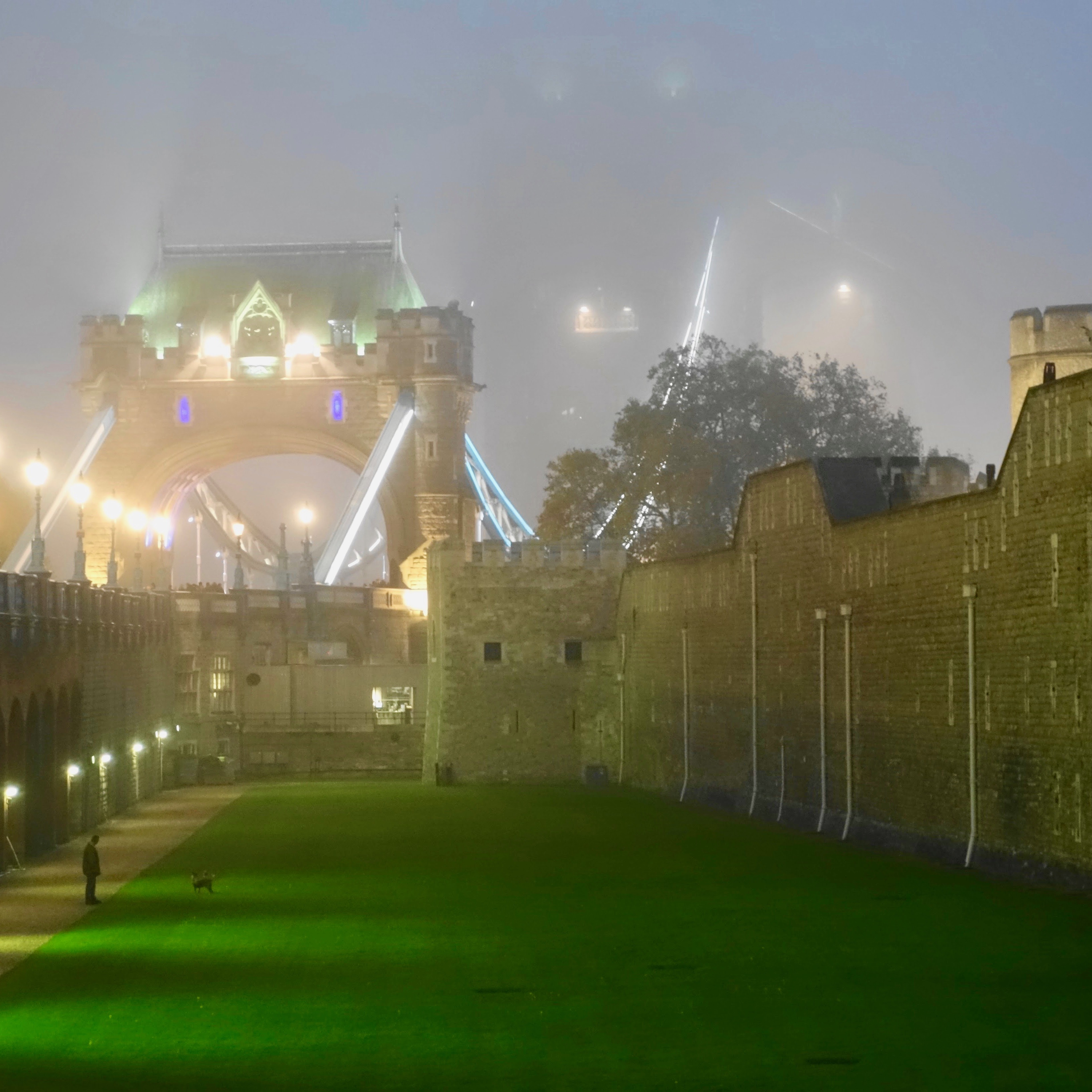 The Tower Bridge in London on a mystic quintessential foggy evening.  The green turrets at the top of the bridge can barely be seen through the fog and to the right lies the famous Tower of London with gray brickwork leading down to a vibrant green grassy lawn.  