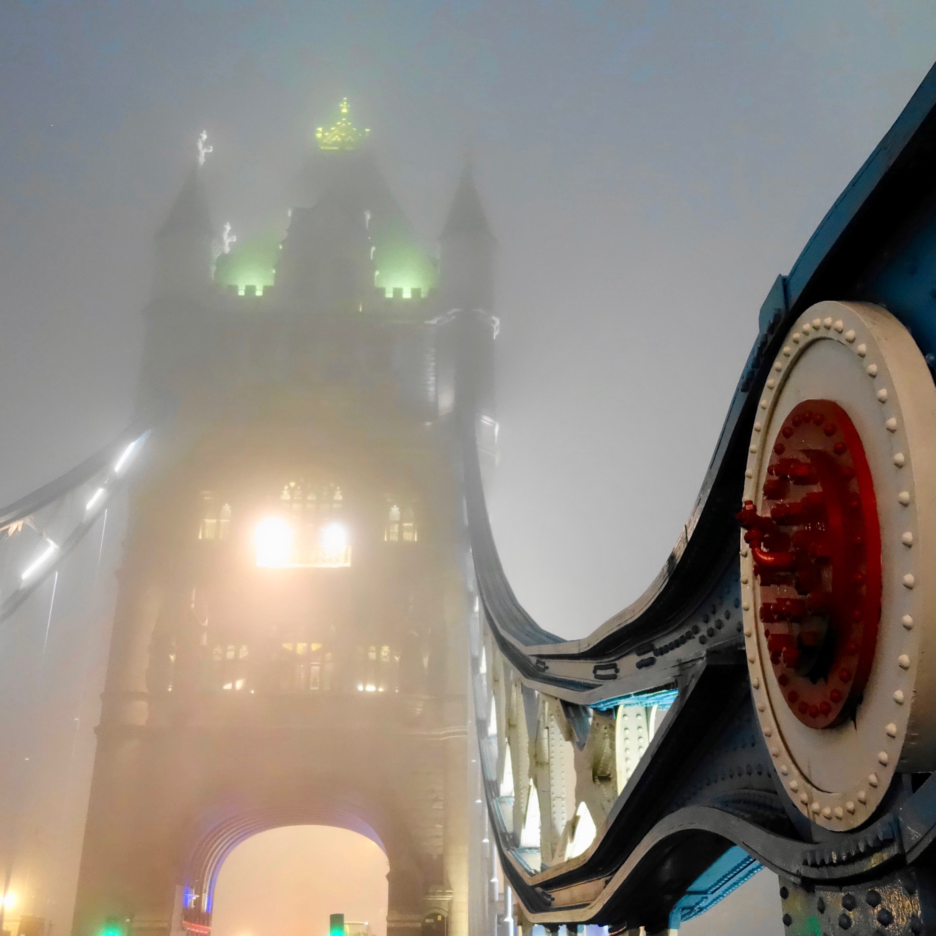 The Tower Bridge in London on a mystic quintessential foggy evening.  The green turrets at the top of the bridge can barely be seen through the fog with a brightly painted red metal structure in the foreground. 
