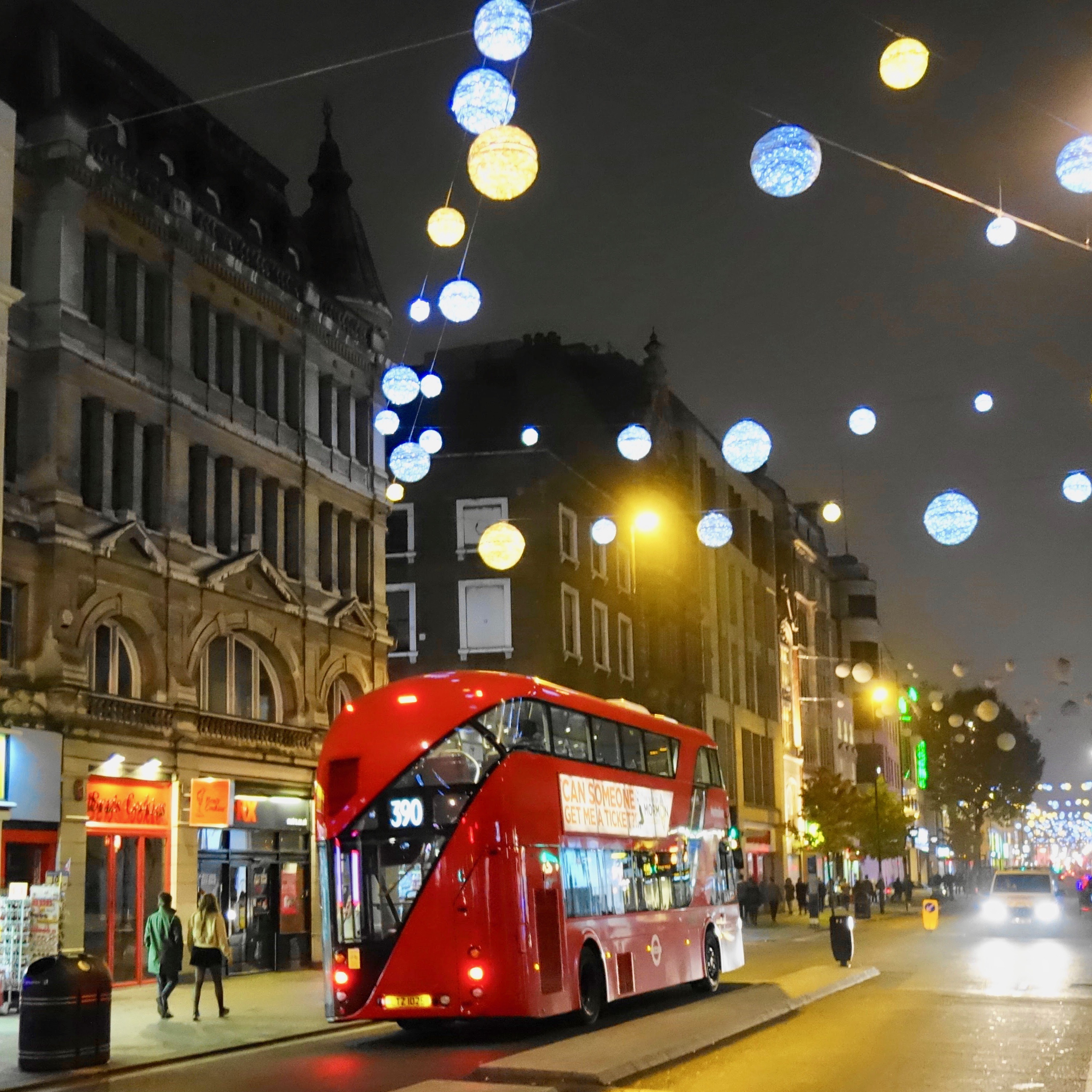 A modern version of a London double decker bus waits for passengers on a quiet street at night.  Holiday lanterns flow across the street high above shining blue tones as a few people walk down the sidewalk. 