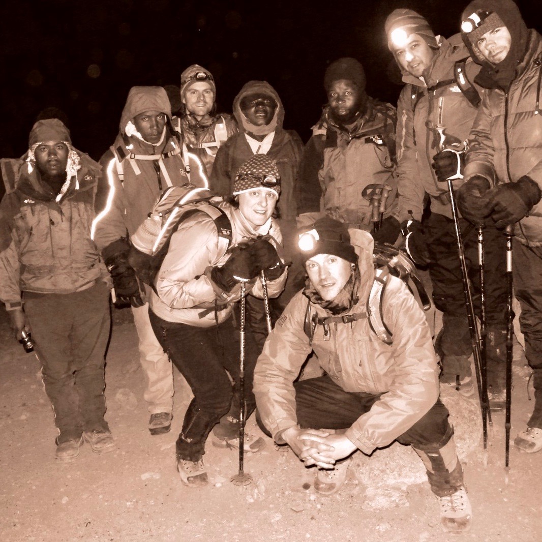 Hikers assemble in the middle of the night, including Matthew Kessi in lower center, ready to climb all night long to the summit of Mt. Kilimanjaro in Africa.  The five climbers have bright headlamps on while the four Tanzanian guides wear reflective parkas. 