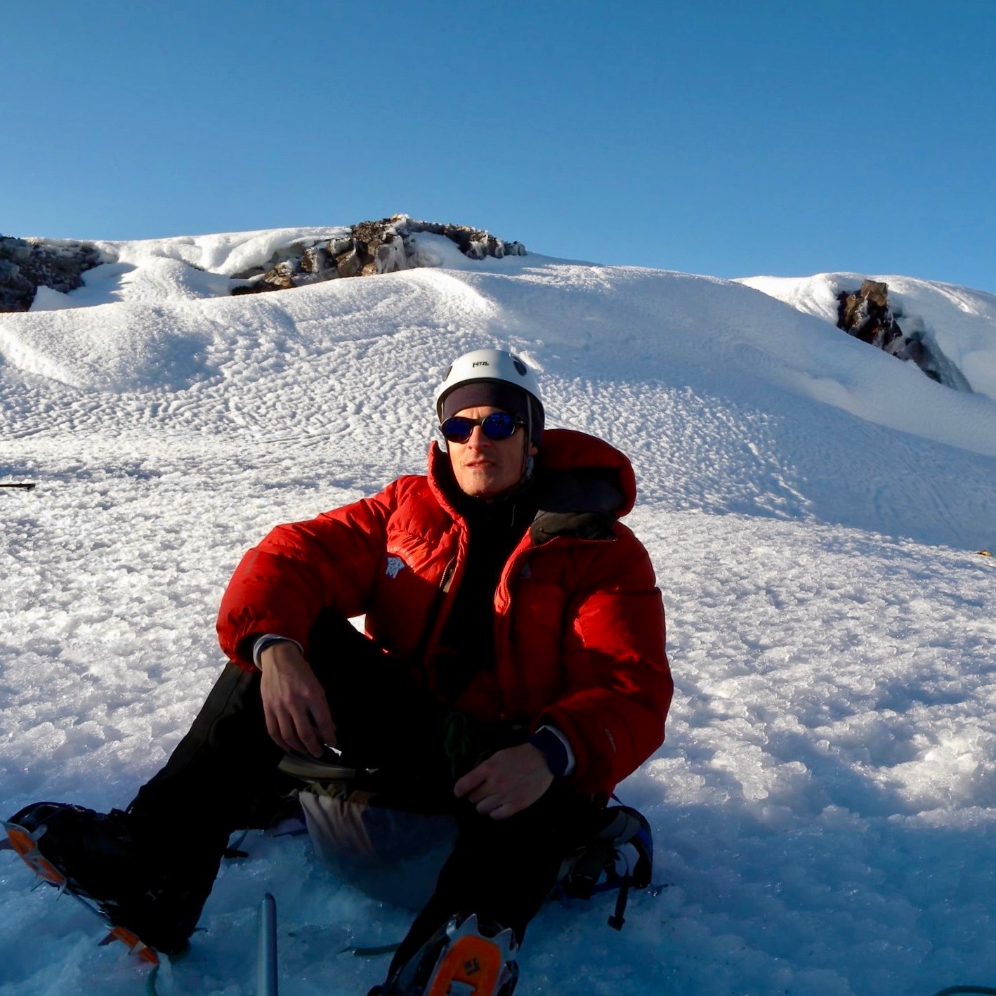 Matthew Kessi sits down on the snow covered glacier at the summit of Mt. Rainier after an all night climb up the side of the mountain.  He's wearing sunglasses and a whit helmet and spikes on the hiking boots.  