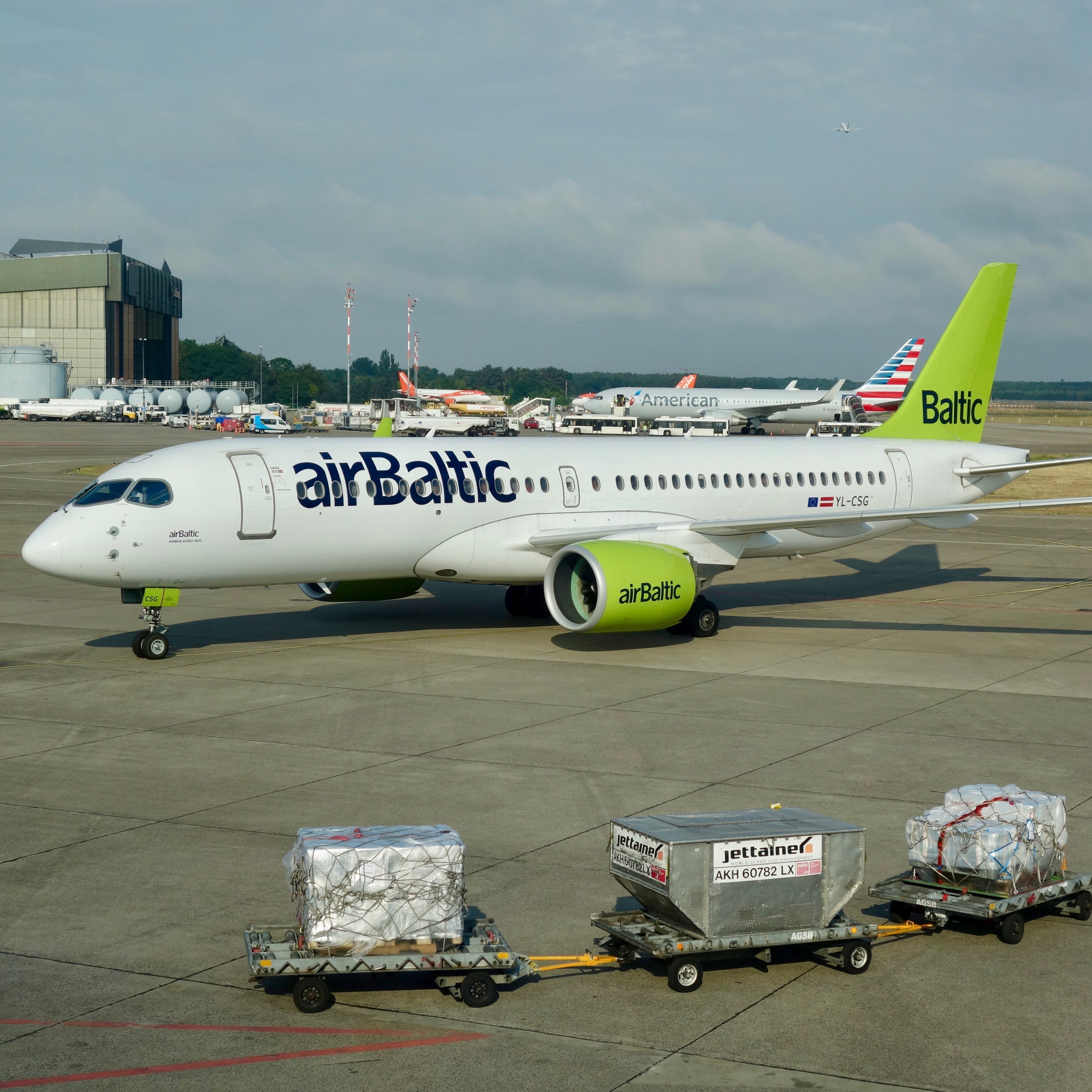 An airBaltic Airbus 220 pulls into the gate at Berlin Tegel Airport while an American Airlines 767 receives servicing on the airport apron.  The new model of aircraft shines with the bright green engines and tail and prominent blue airline logo on the forward fuselage.  This review of the experience reveals an efficient airline with modern services. 