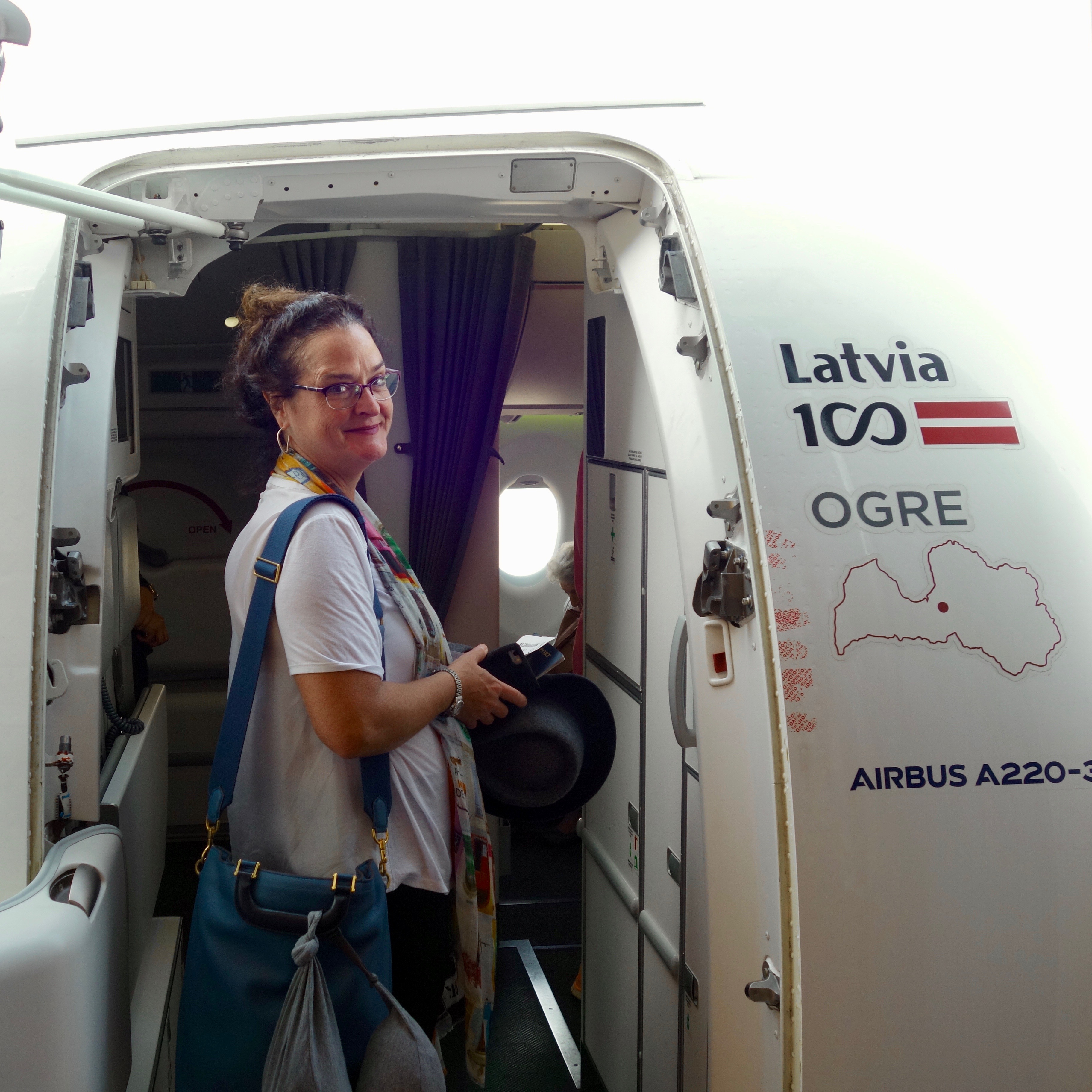 A female passenger boards through the front main cabin door of an airBaltic Airbus A220.  The outside of the fuselage shows a few different stickers highlighting the airframe type and a map of Latvia.  