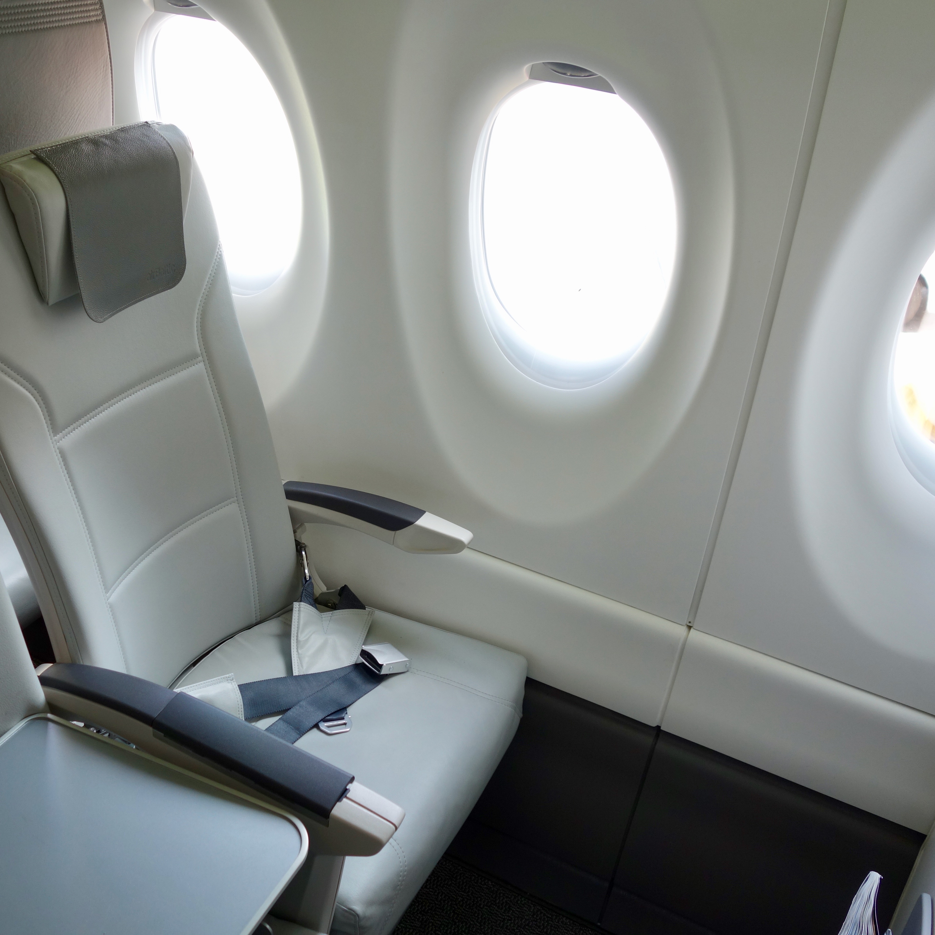 First Class section of the airBaltic Airbus A220 shows regular coach seats with a blocked seat next to it to allow for more space.  the seat is made from leather and the large windows allow in abundant light. 