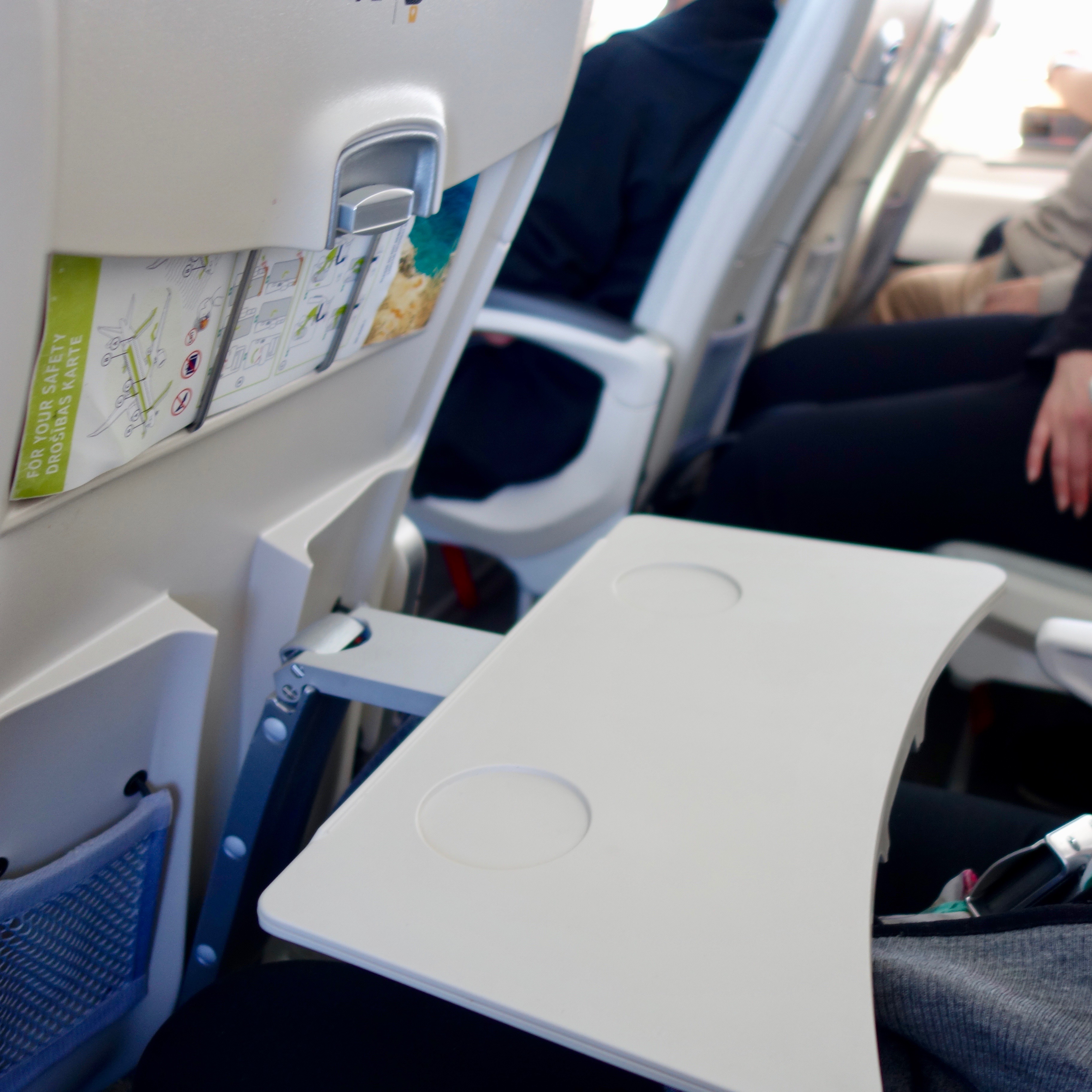 The tray table of an airBaltic A220 is beige colored and utilizes a unique metal bar, which is a new design. 