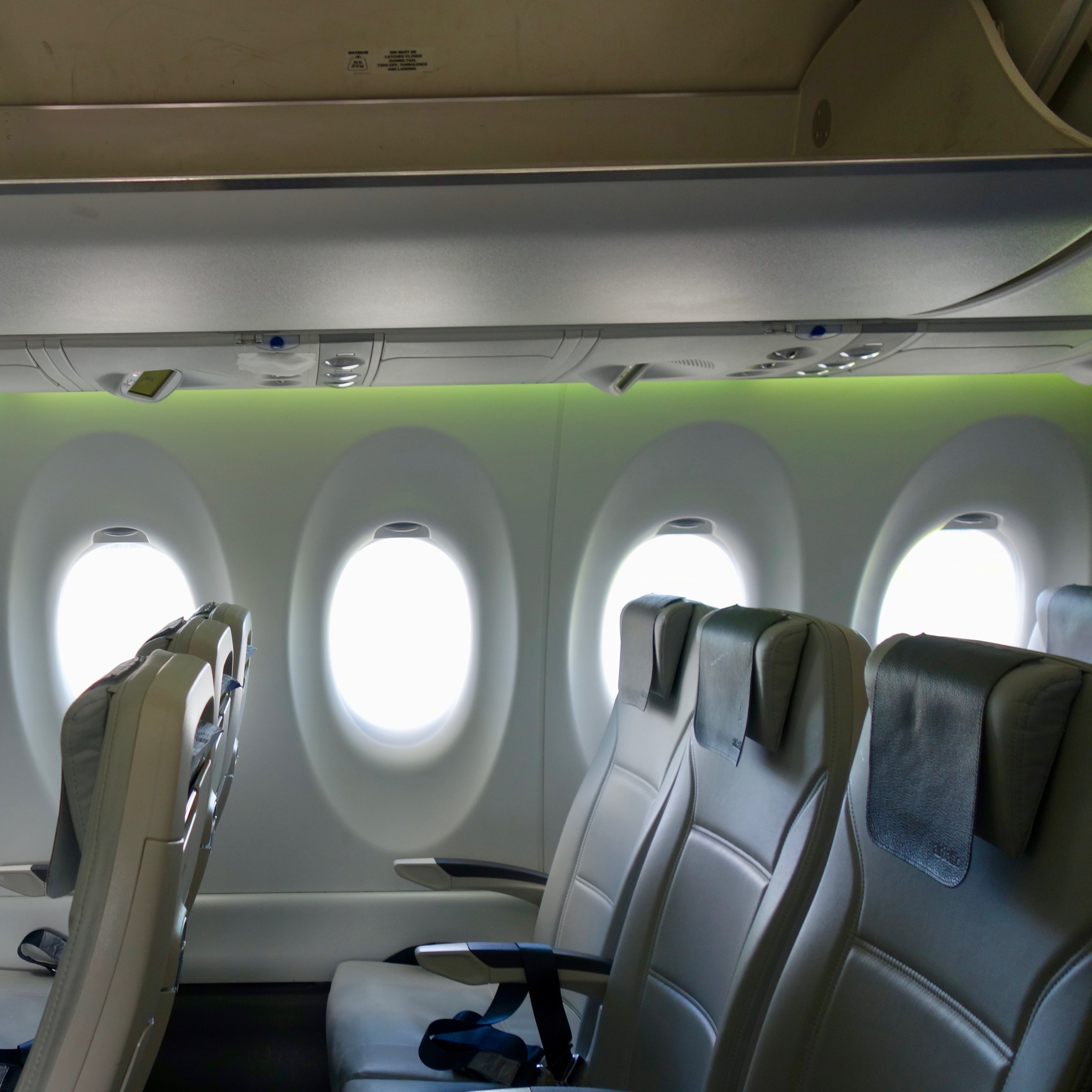 The Airbus A220 is a modern airliner, and airBaltic adds their branding touch with green neon cabin lighting shown adjacent to the overhead luggage bins.  The three seats leading to the large sunny windows are made from gray leather. 