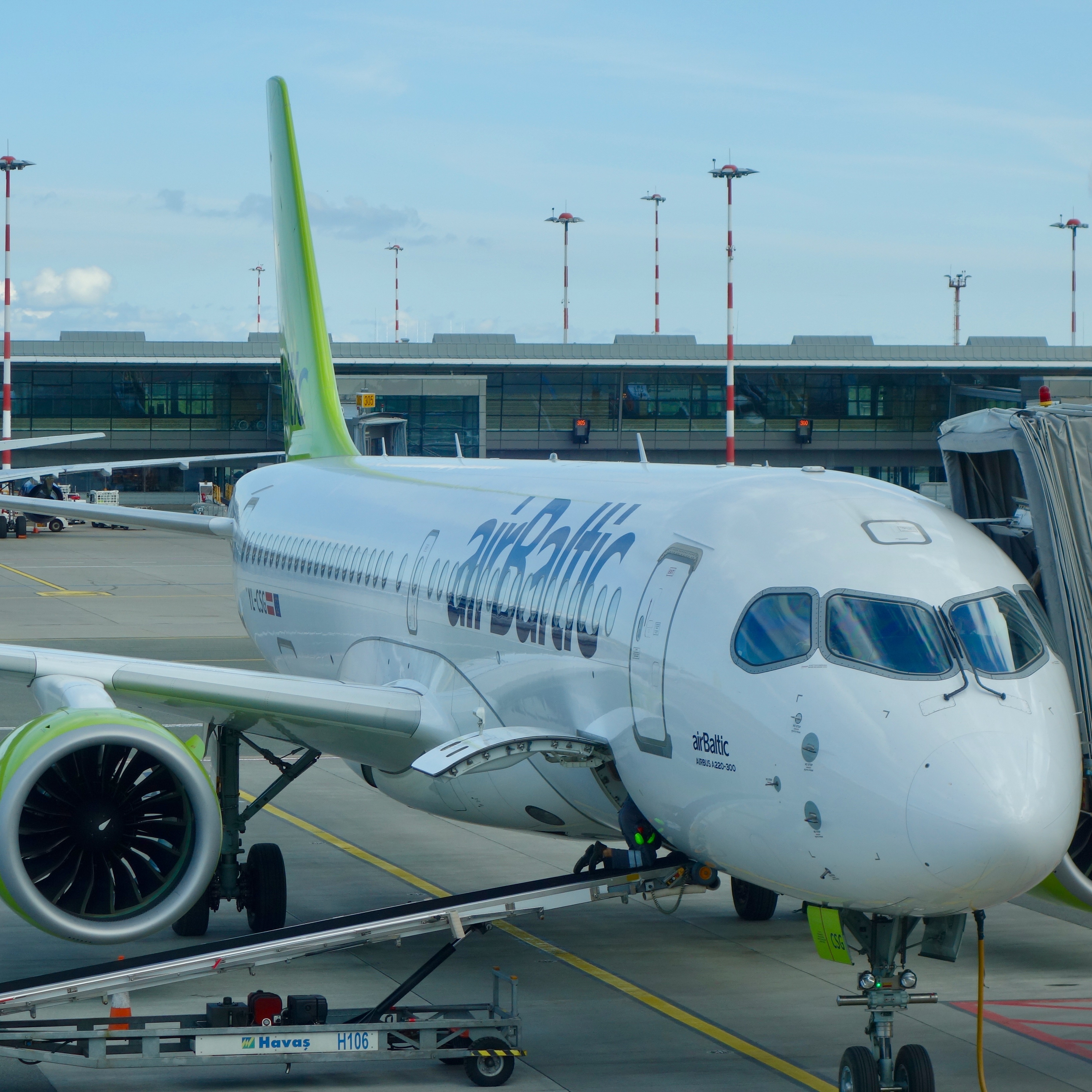 The modern, sleek look of an airBaltic A220 waits at the gate for passengers to board in Riga, Latvia.  The flight deck windows are oversized and shiny turbines of the round engine temporarily quiet.  