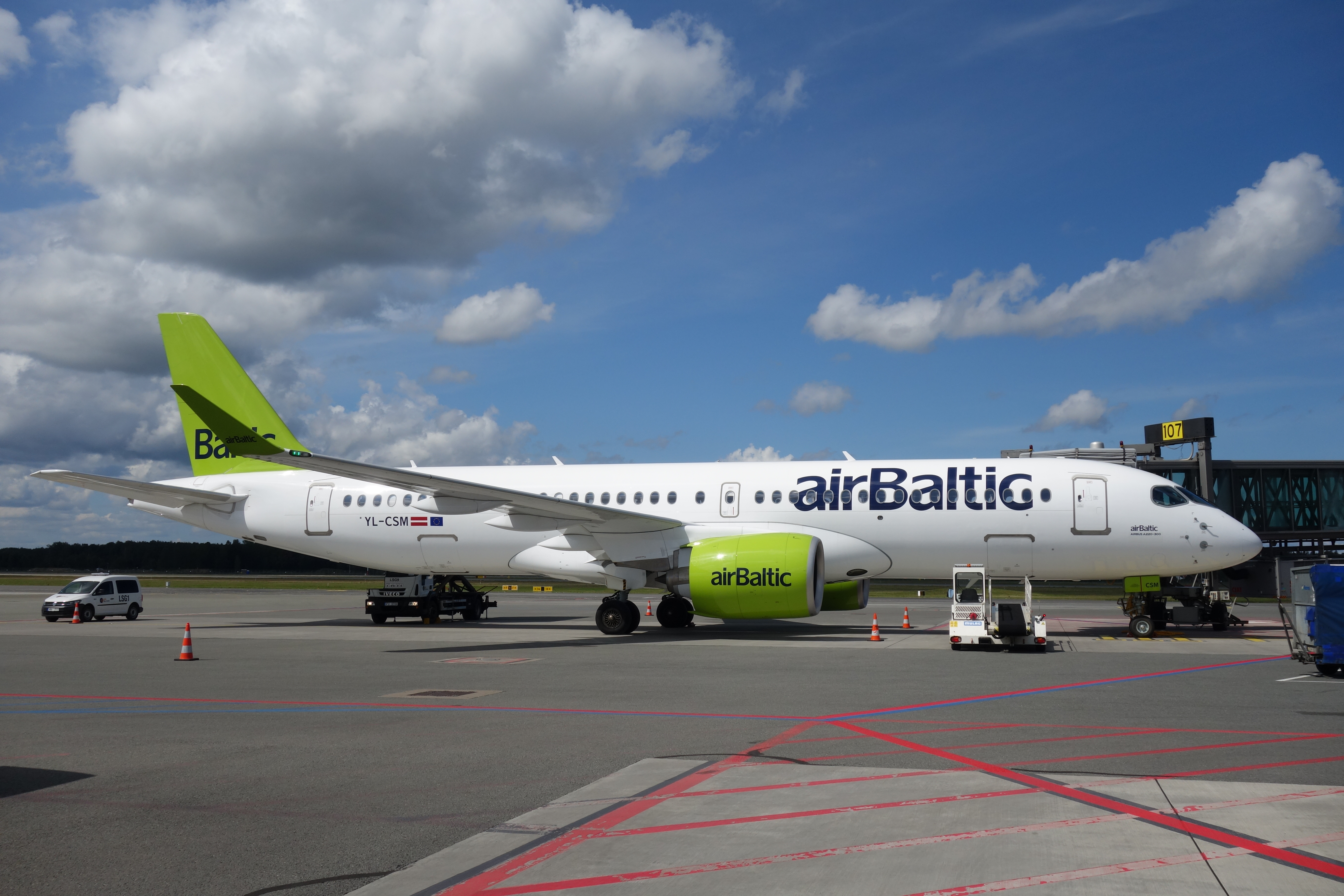 A brightly painted airBaltic Airbus A220 prepares for departure at a gate in Riga, Latvia.  The engine and tail is a chartreuse green while the fuselage is white and the airline name printed in navy blue. 