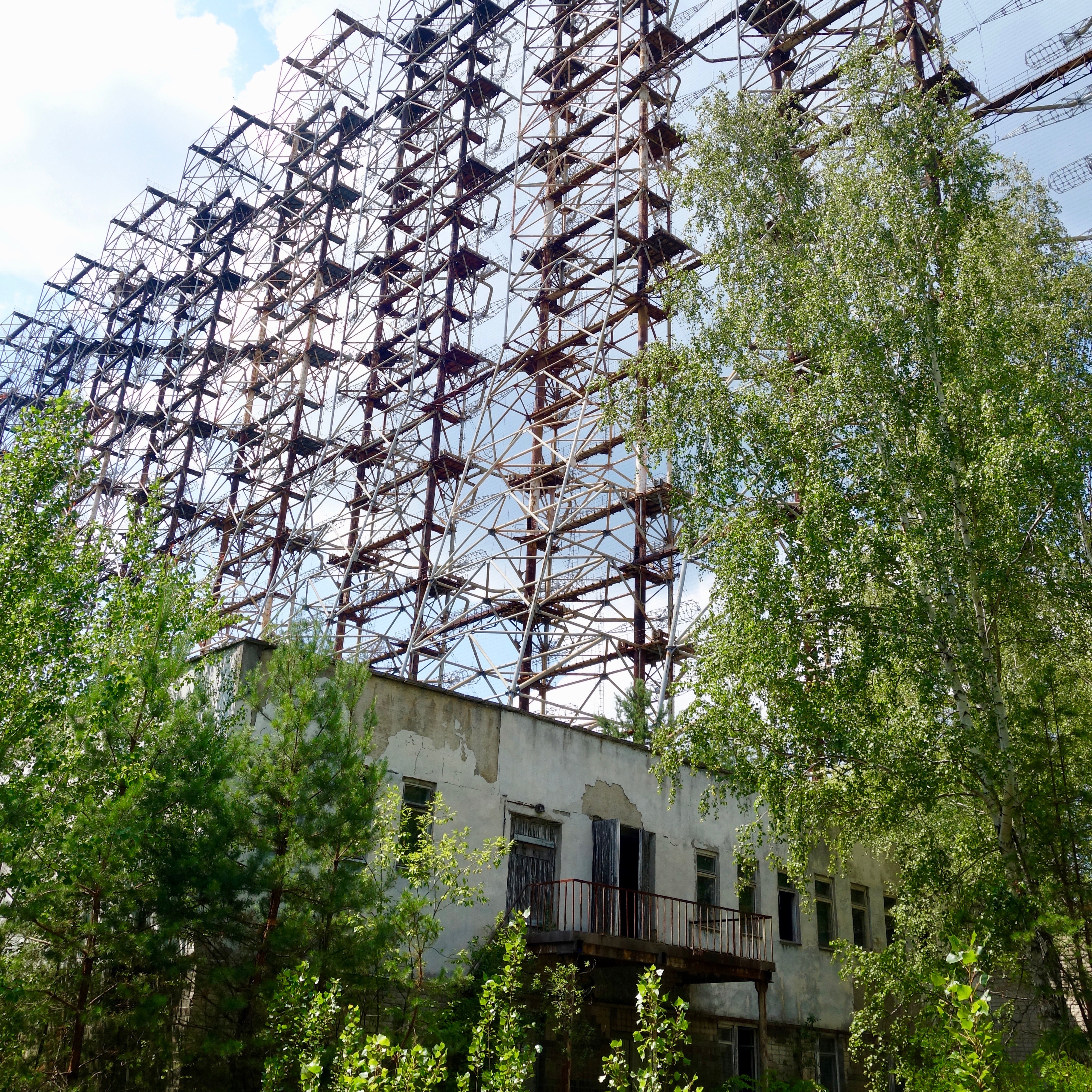 The Russian Woodpecker was a giant structure of metal attennae like structure rising up from a pine tree forest only a few miles from the Chernobyl Reactors.  This shot shows the giant rusted metal structure rising up to the blue sky with green willows amid other smaller green pine trees with reddish brown tree trunks.  