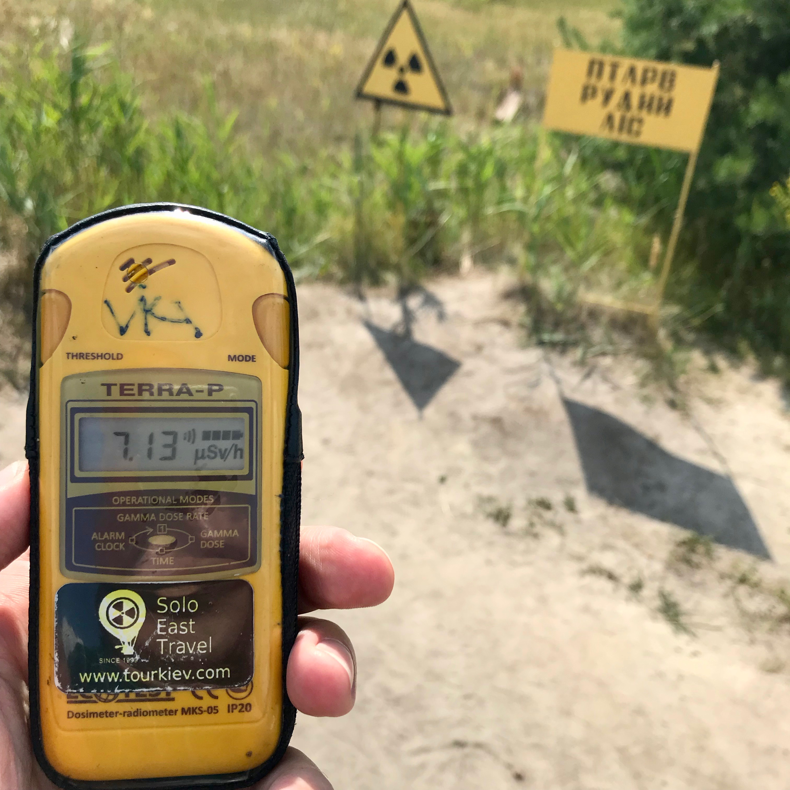 A yellow Geiger counter with the level of radiation in the area, which in this case is displayed at 7.13.  I'm holding this device above an area that was used to bury all the radioactive trees of the forest.  The ground is a dusty brown with a field of grasses in the periphery.  