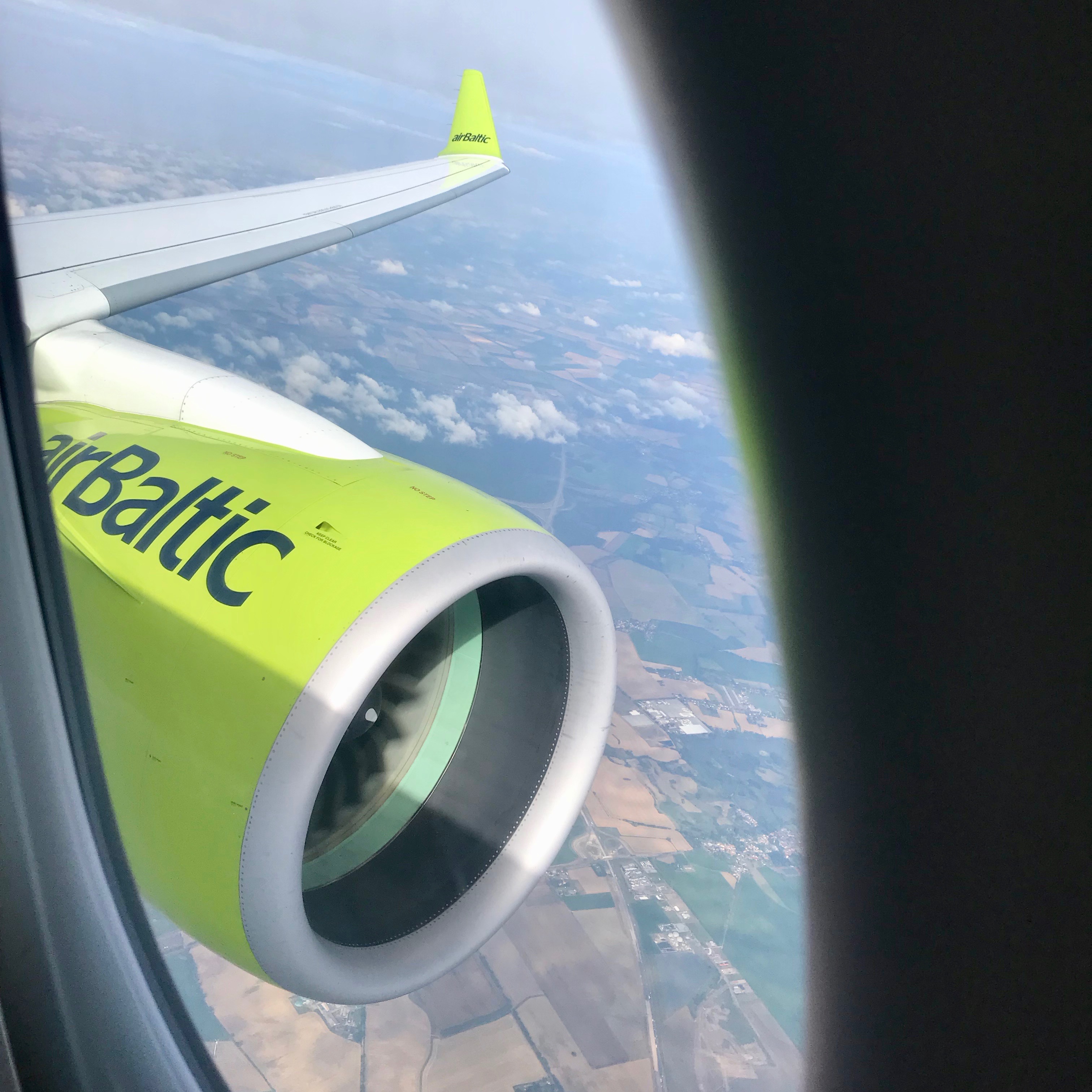 A view out the window of an airBaltic Airbus A220 shows a bright green engine with the airline logo prominently displayed while a green wing tip pops up.  The jet is flying high above the clouds. 