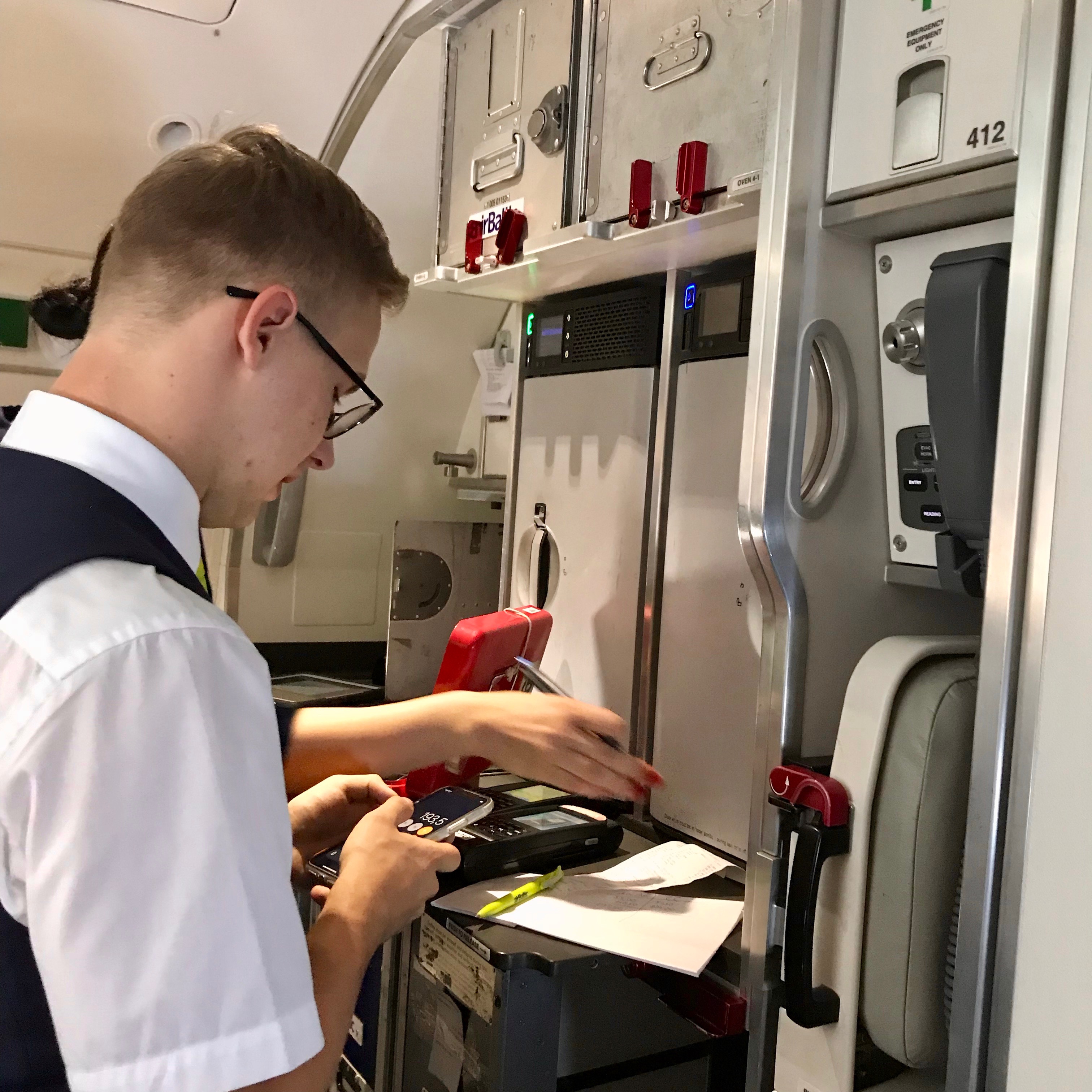 A flight attendant in the aft cabin near the galley of the airBaltic A220 conducts accounting duties with an iphone.  The review of the airline experience revealed friendly crews. 