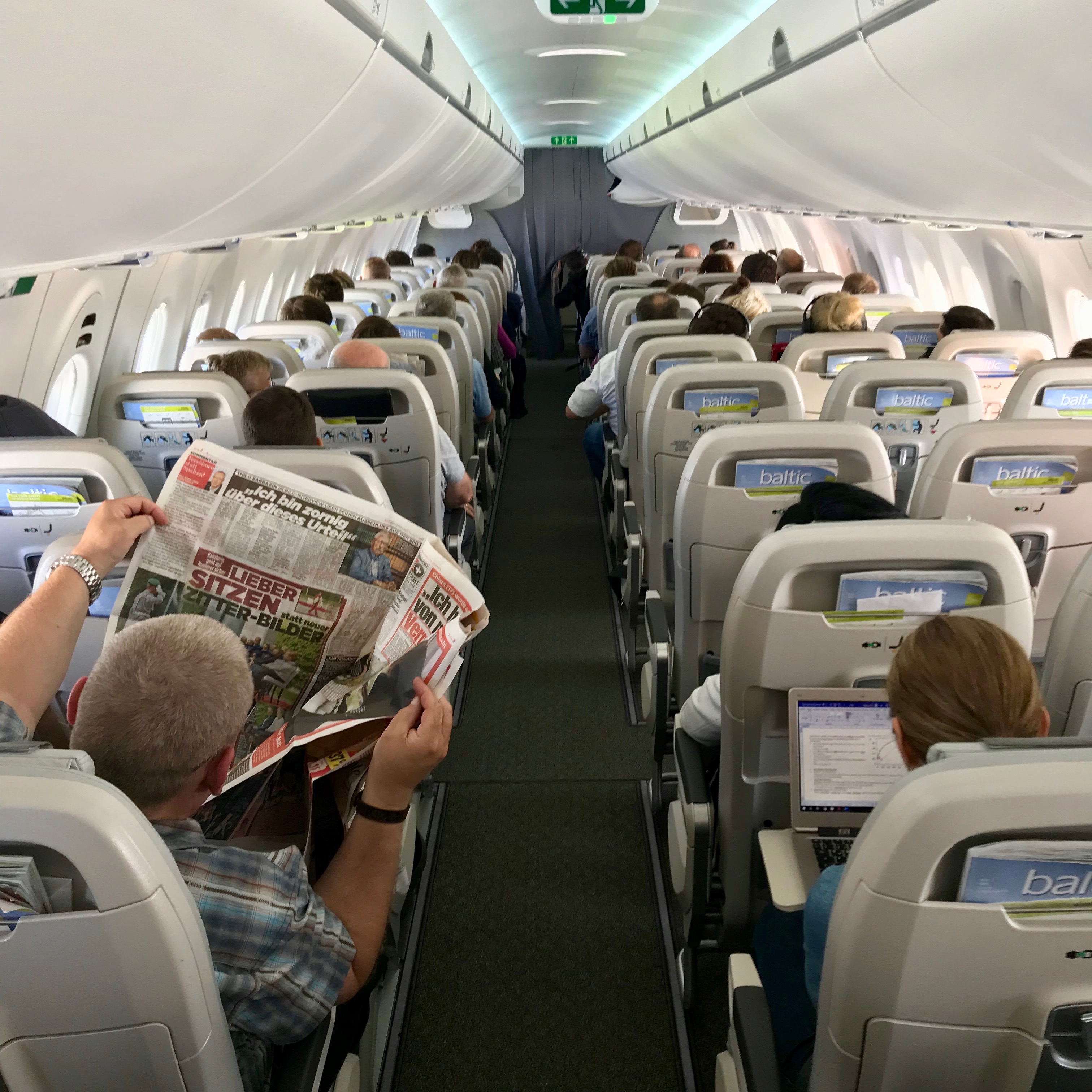 The inside view of an airBaltic Airbus A220, from the aft cabin reveals a five across seat plan.  Passengers read newspapers and work on laptop computers while the large open windows allow in lots of light. 