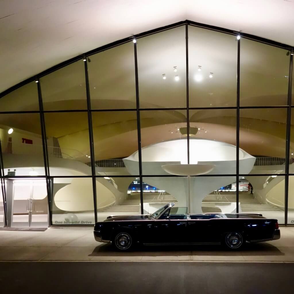 View from the front of the TWA Hotel looking into the iconic 1962 terminal. In front of the wall of windows is a 1960's vintage Lincoln convertible.