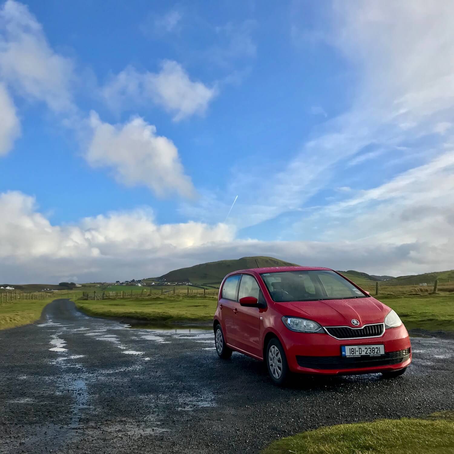 Red rental car on a county road in County Donegal, Ireland. Grassy hills and blue skies frame in the farmland.