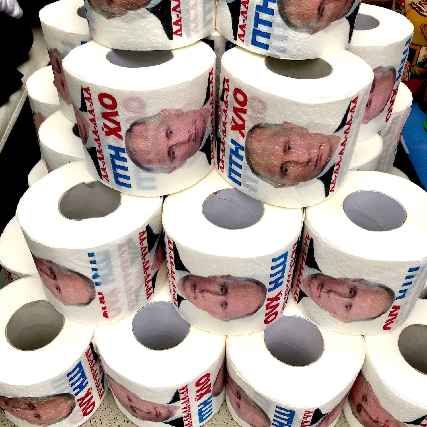 Items for sale at a street side vendor booth.  Here is a pyramid of toilet paper with Vladimir Putin's face. 