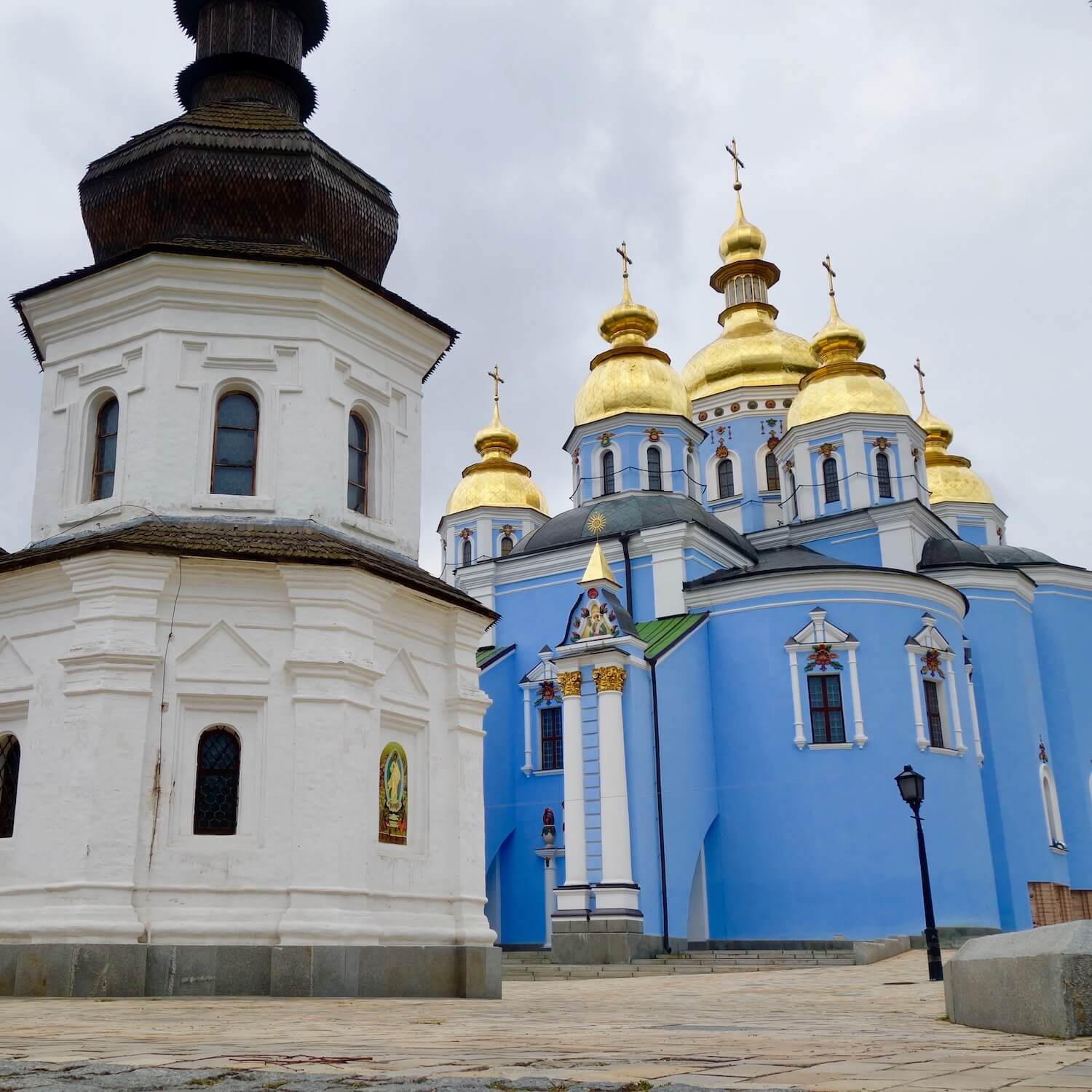 St. Michaels Monastery is a complex of orthodox buildings including the older wood church with white walls and black onion dome in the foreground and the bright blue main church with golden spires in the background.