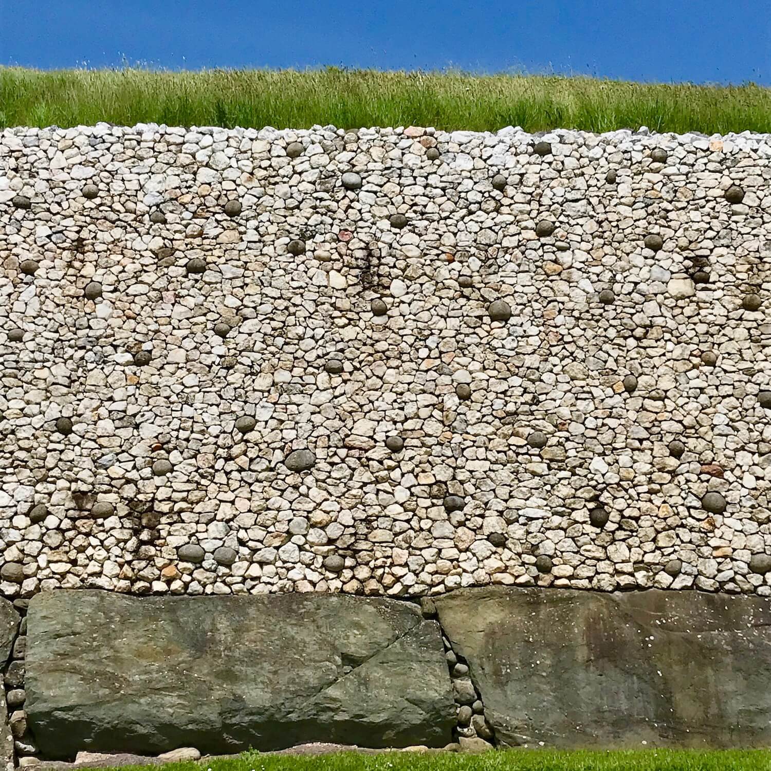 A closeup look at the Newgrange mound shows a wall of whitish stones with larger gray ones interspersed. The wall is framed by green grass on the top of the mound and large granite stones forming a base on the bottom.