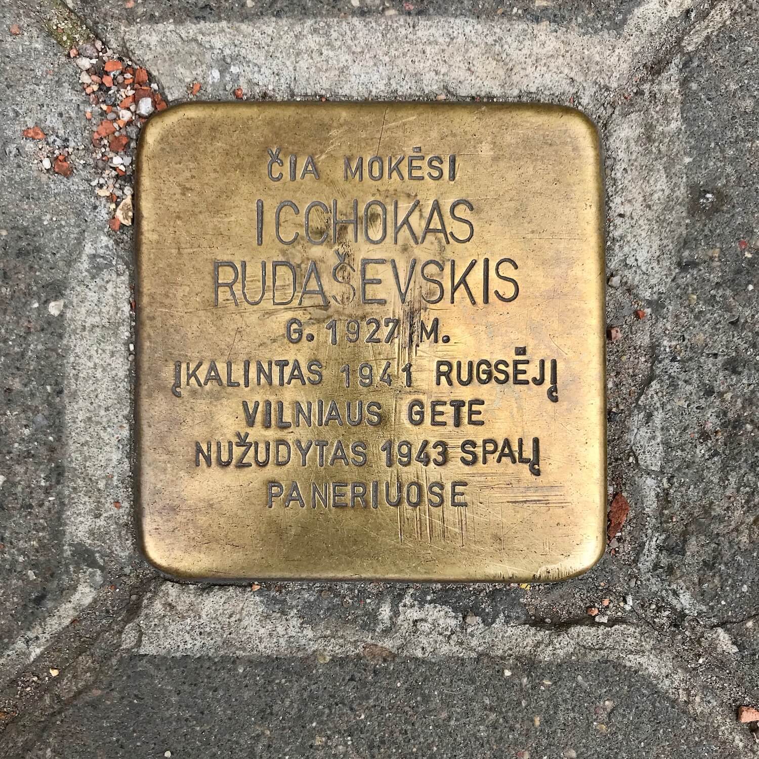 A copper tile on the sidewalk in Vilnius Lithuania remembered a Jewish citizen taken from their home and sent to concentration camps to die. The writing is in Lithuanian.