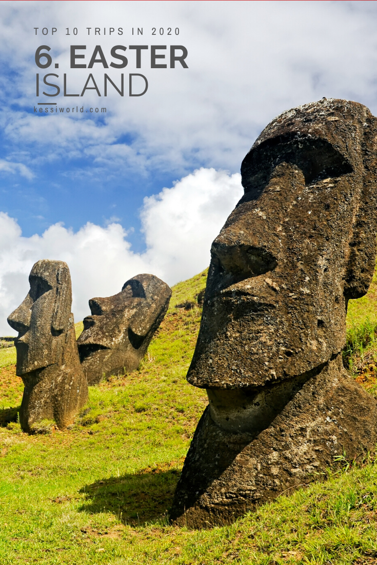 This shot of Easter Island shows a prominent rock statue with a primitive looking face amongst a grassy green hillside. In the background there are two other similar busts of primitive looking men and the sky is blue with some clouds.