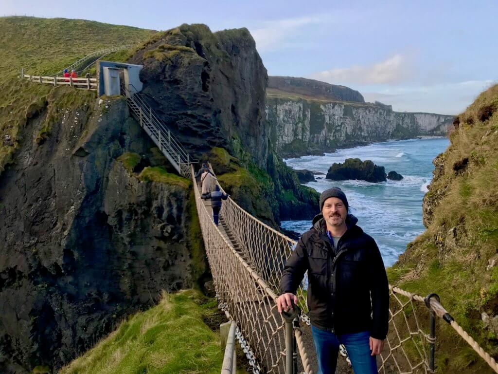 This photo shows the Carrick-a-Rede rope bridge, which spans from a rocky cliff along the Atrim coast to a small island. Above the rock base are rolling waves of green grass. Matthew Kessi stands near the bridge as an example of how to nature forward on vacations.