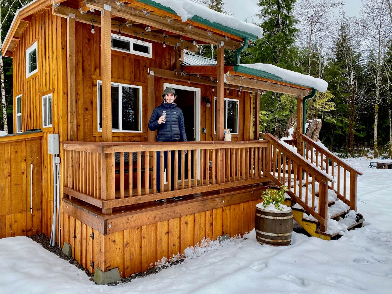 The exterior of a tiny house with Konstantin sipping coffee on the front porch. The cabin has six inches of snow on the roof lines and wet pine siding in a western style.