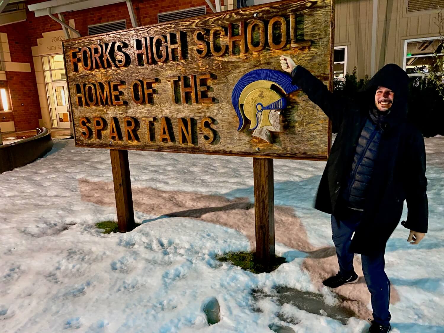A man stands in front of a sign that reads "Forks High School, home of the Spartans". There is snow on the ground and the school building in the background.