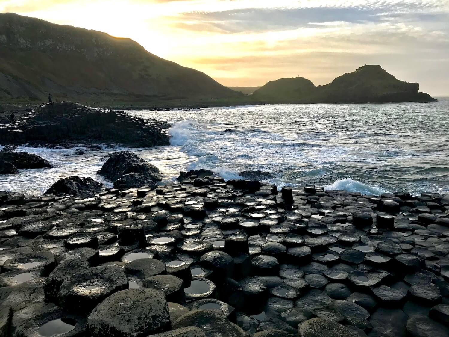 The Giant's Causeway is an area of about 40,000 interlocking basalt columns which are shown in the foreground of this photo. Dramatic high cliffs leading to the sea are in the background with blue sky.
