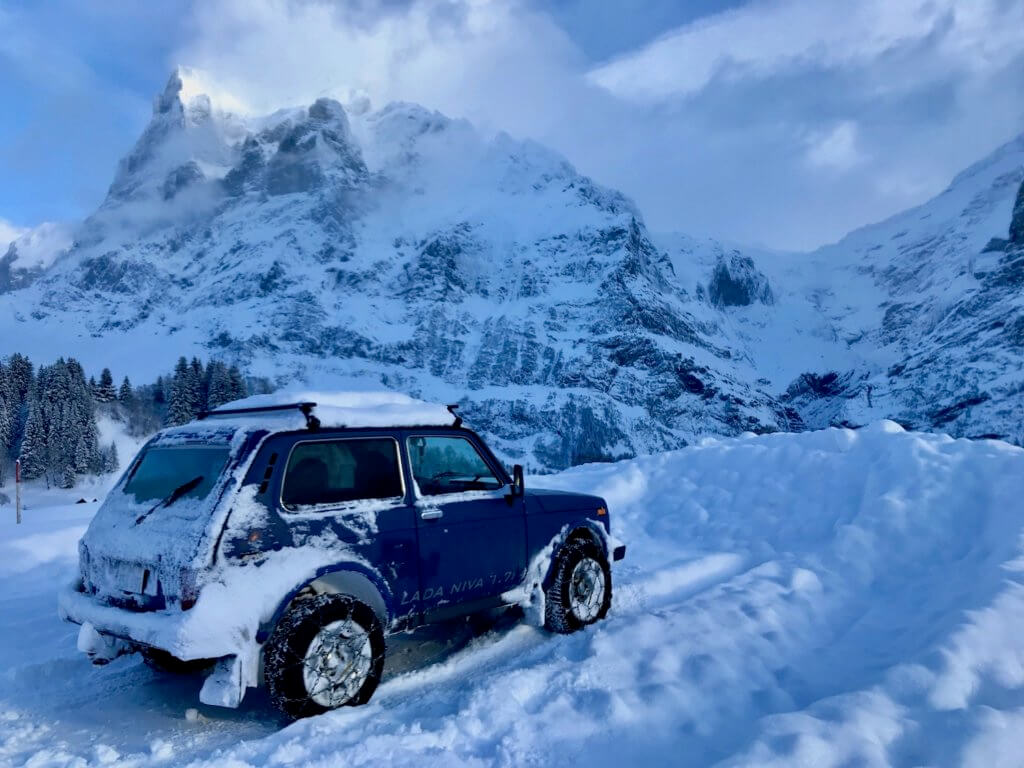 A tight little blue hatchback vehicle with chains on the wheels sits in a deeply snowy car park, the owner likely out snow shoeing or hiking. In the background are high alpine peaks. This is near Grindelwald Switzerland in a snowy winter scape.