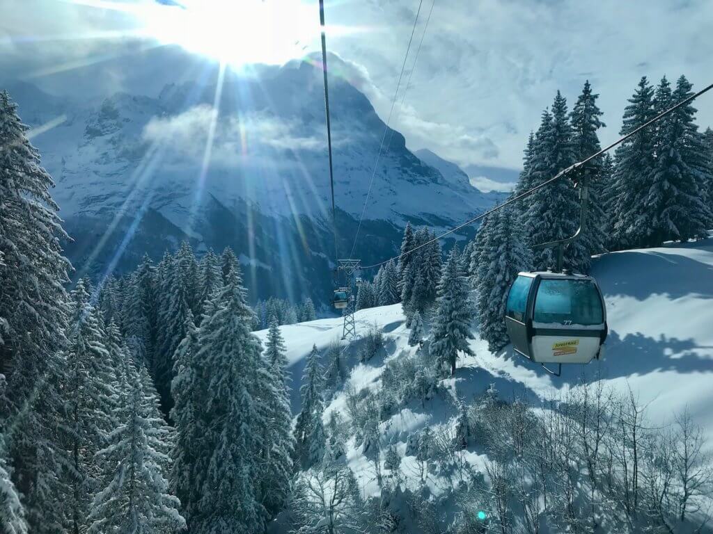 View of the sun shining over the top of an alpine peak near Grindelwald, Switzerland as the ski gondola pulls up the mountain. Below the gondalas are snow covered fir trees and uneven snowy terrain.