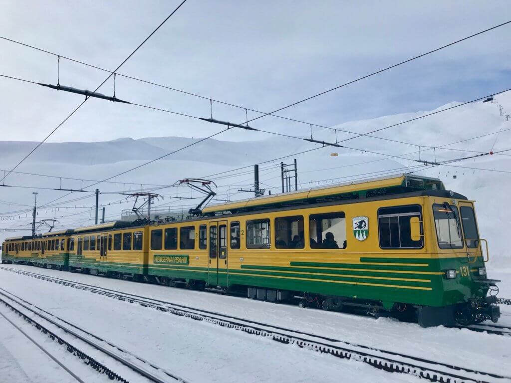 A shiny green and yellow painted Swiss train in the rail connection hub of Kleine Scheidigg. The narrow gauge rails are popping up through the snow as well as several electric power lines, driving the rails.