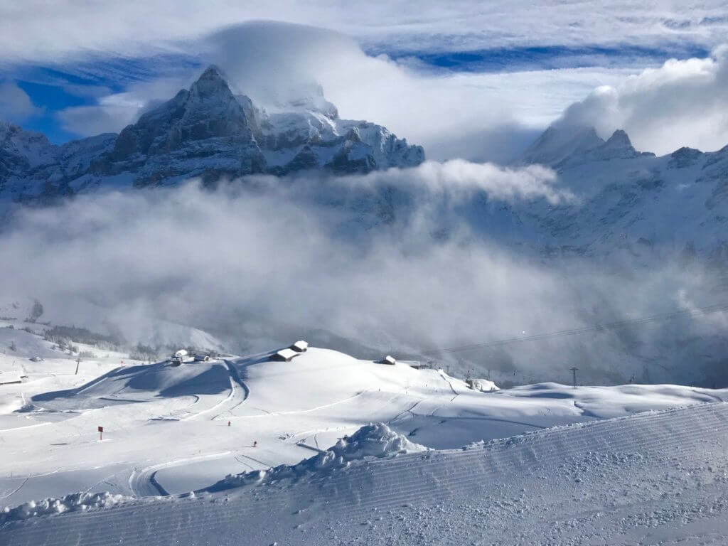 View from a ski trail overlooking groomed ski runs with snow covered chalets leading down the mountain. In the background are the dramatic Alpine peaks of Schreckfeld and Schrekhorn. The clouds surround the peaks in a dramatic way that opens to some patches of blue sky.