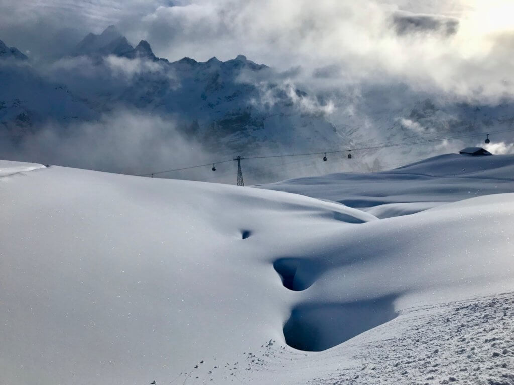 View of the glistening snow from a groomed ski trail in the Swiss Alps near Grindelwald. In the background is a ski gondola and the sharp snowy peak of mountains in the background. The mountains are steaming with active looking clouds.