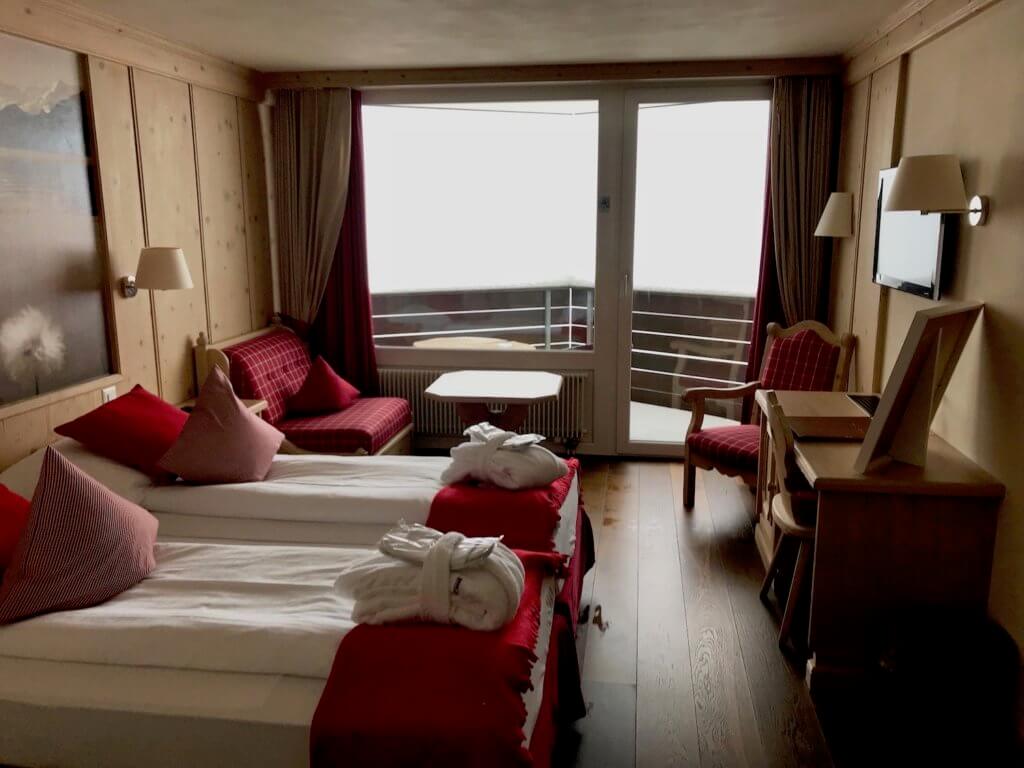 Hotel Spinne room with two full beds next to each other and red splashes of color with pillows, blankets and chair coverings. The balcony looks out to a snowy blizzard outside.