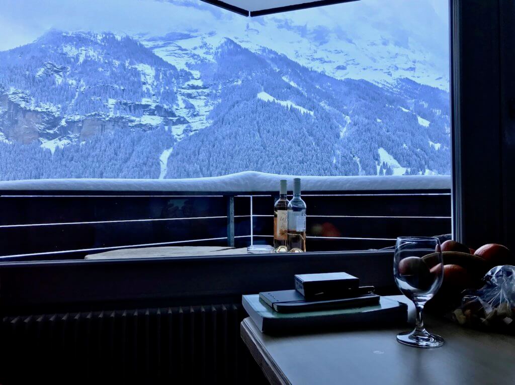 View from a hotel room at Hotel Spinne looking past a table with fruit and books to the balcony table with two bottles of wine just beneath the rail with four inches of snow. In the background is the base of Eiger (13,000ft).