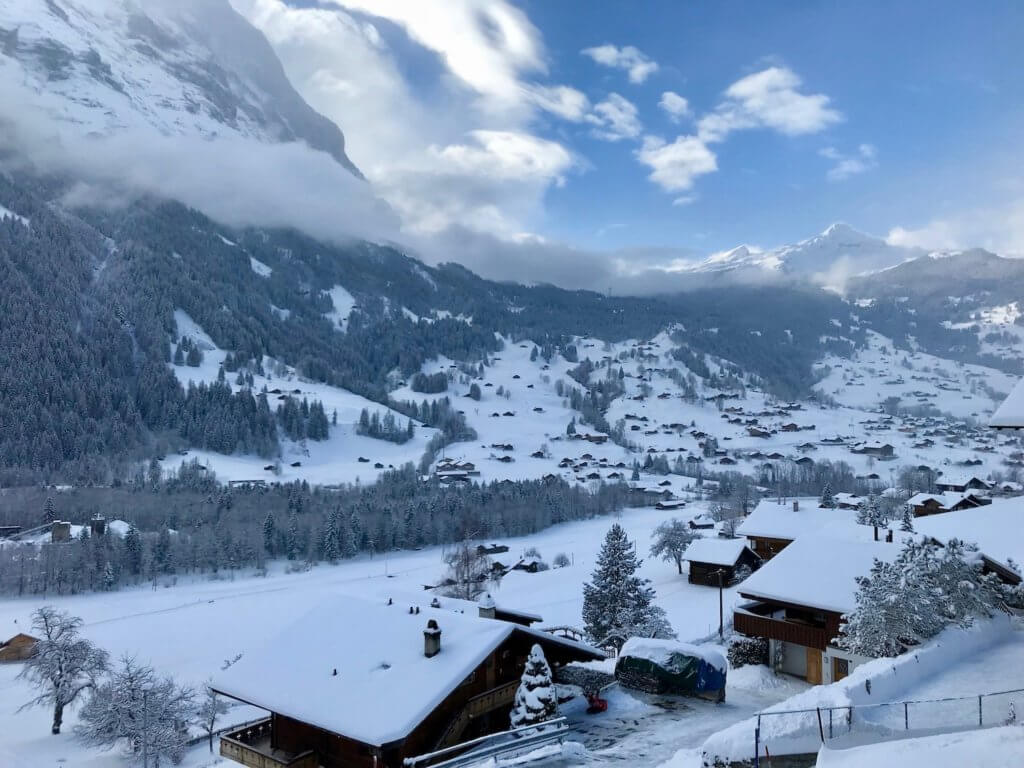 View from a hotel balcony in Grindelwald viewing the gentle hillside with snow covered chalets and two high alpine peaks in the background hugged by light puffy clouds and blue sky on the top of the photo.