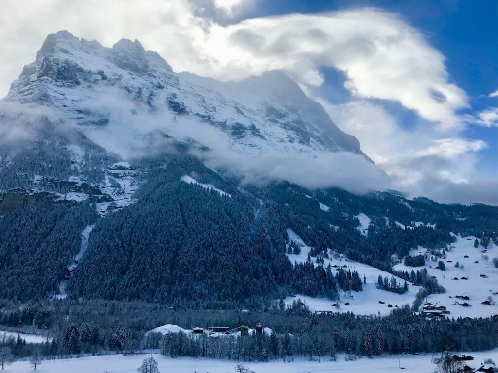 View from Hotel Spinne in Grindelwald, Switzerland towards the dramatic snow covered Eiger (13,000) feet. The sky is a mix of bright blue with a swirl of fluffy white clouds.