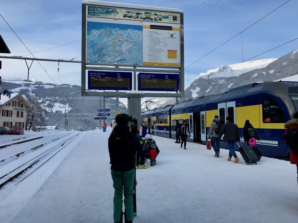 The main train statin in downtown Grindelwald. The blue and yellow regional train looks ready to head to Interlaken as passengers wheel their luggage to board. A woman in the foreground glances up and the large map of the mountain terrain and train information monitors. The ground is snowy and mountain peaks with blue sky in the background.