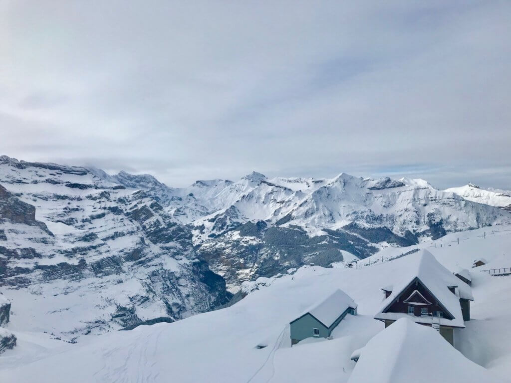 View from the rail platform at Eigergletscher looking out towards numerous snow covered alpine peaks. In the foreground are three snow-covered Swiss chalets the hovered on the side of a steep ski slope.
