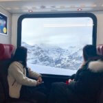 View from the narrow gauge rail train leading up to Top of Europe tourist attraction. Two asian tourists peer out the window of the train to a dramatic landscape of jagged Alpine peaks.