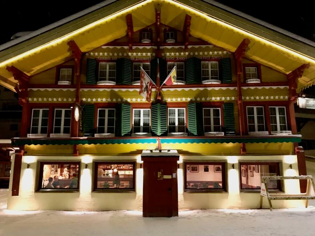 A colorful restaurant in traditional Swiss architecture style with bright yellow eaves and walls contrasting to greens shutters and red timbers. This shot is an night and the bright streetlights shine on the snow covered road.