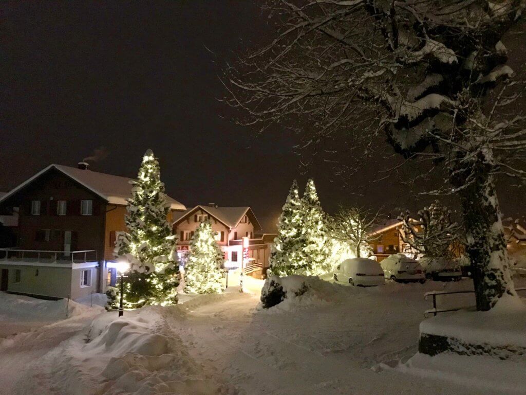 The road leading into downtown Grindelwald is ablaze with lit up Christmas trees and other festive lights. There are several snow covered chalets and a few snowy cars in a parking area.