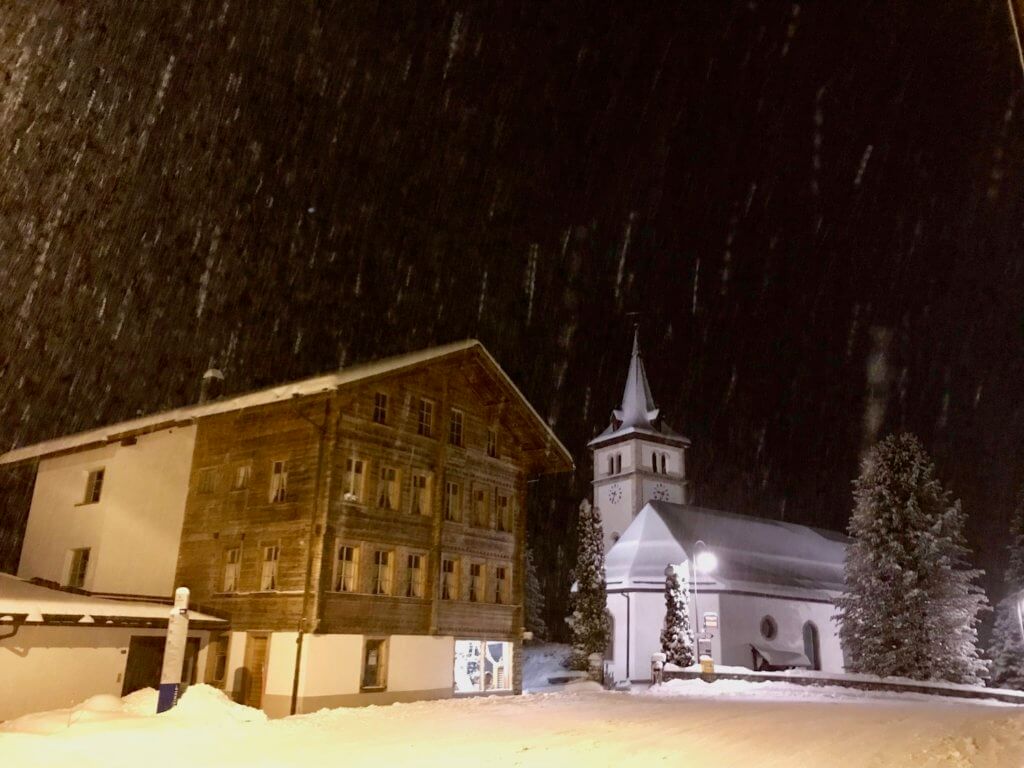 Downtown Grindelwald at night with the falling snow streaking through the photo. A wood structured guest house and the town church are flooded with light from the street as the snow falls.