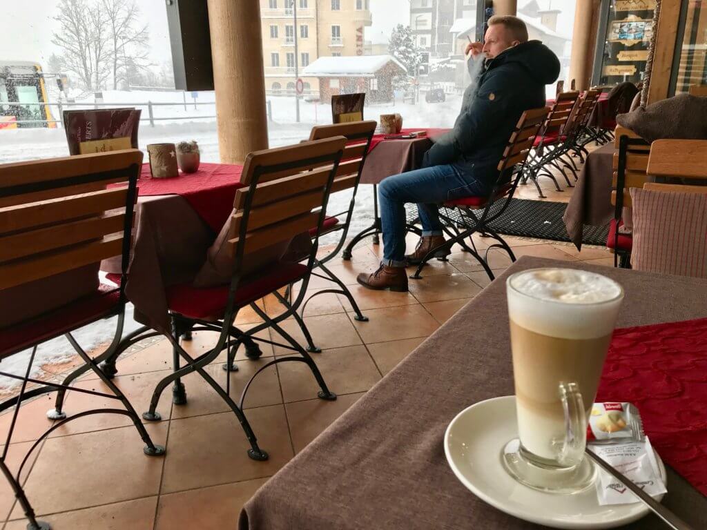 Snack Bar Kreuz & Post is a street side cafe with bistro chairs and tables covered with red tablecloths facing the snowy street. In the distance a man in a Canada goose parka smokes a cigarette and looks on. There is a cafe latte at the table in the foreground with a fluffy milky foam on top of the drink.