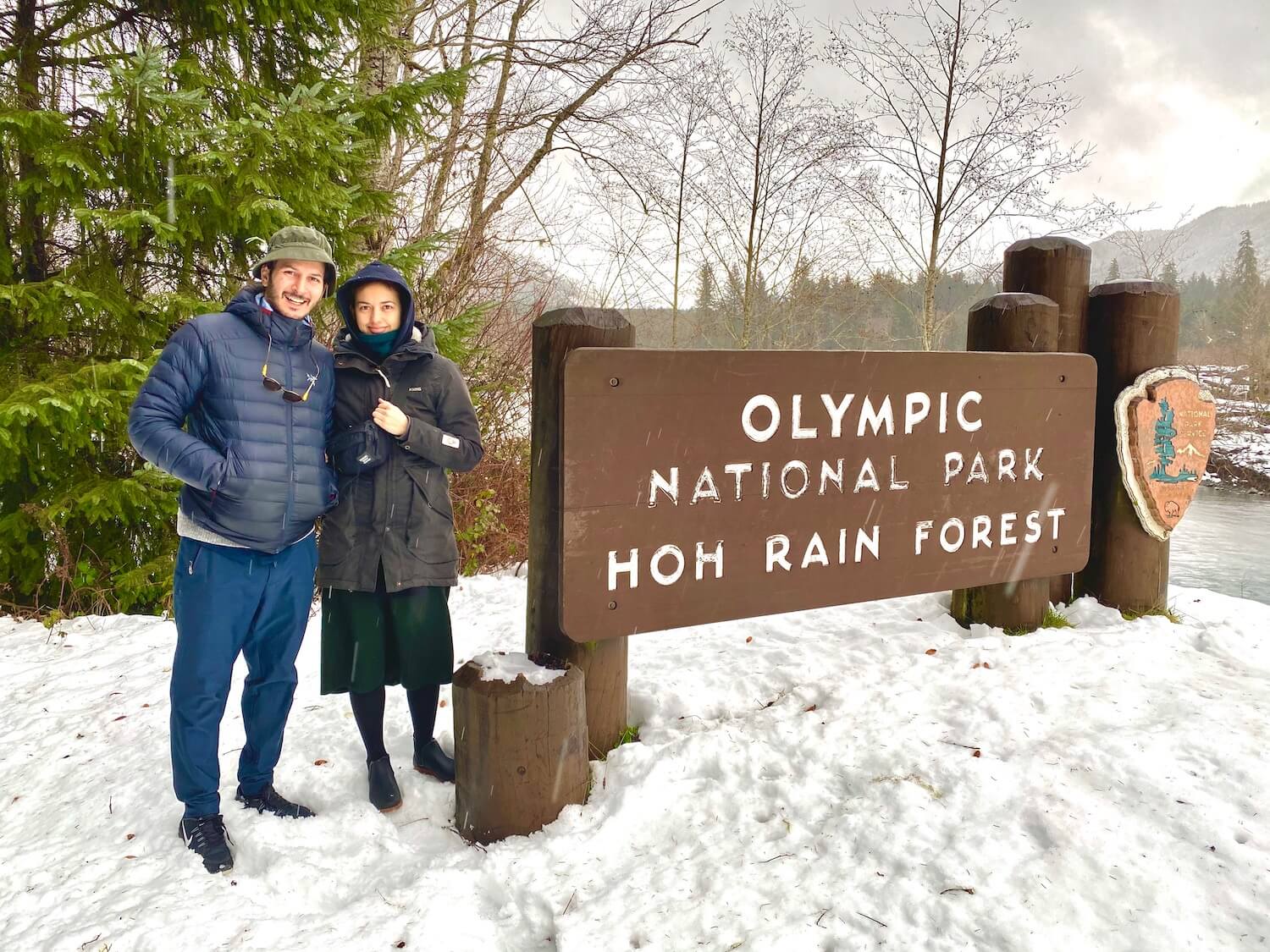 My friends stand at the entrance to the Olympic National Park. There are green fir trees in the background and snow on the ground.