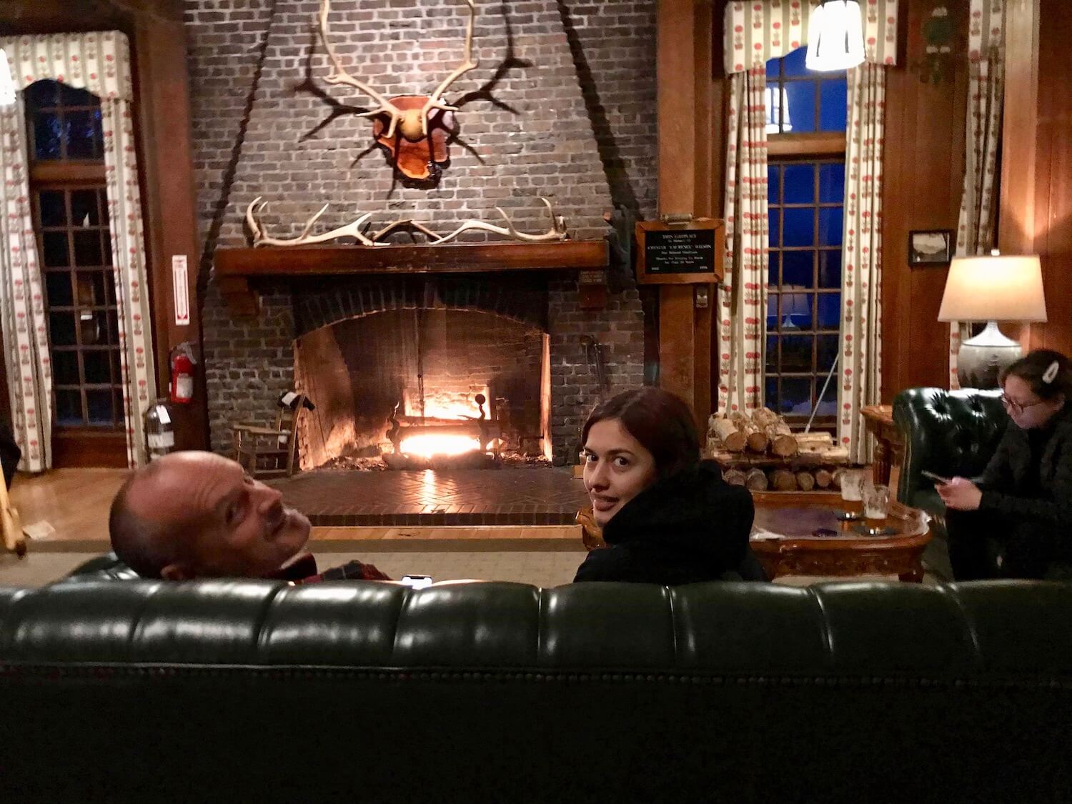 People relaxing on plush green leather couches in front of a grand fireplace inside a lodge. There are Roosevelt Deer antlers on the mantle of the fireplace and also hanging on the stone chimney.