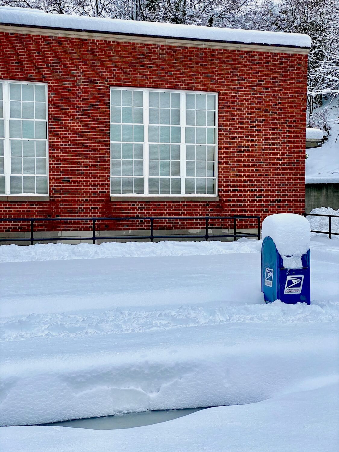 A US post box is covered with a foot of snow precariously hanging on. In the background is the red bricked post office with six inches of snow on the roof. The foreground has a two feet of snow.