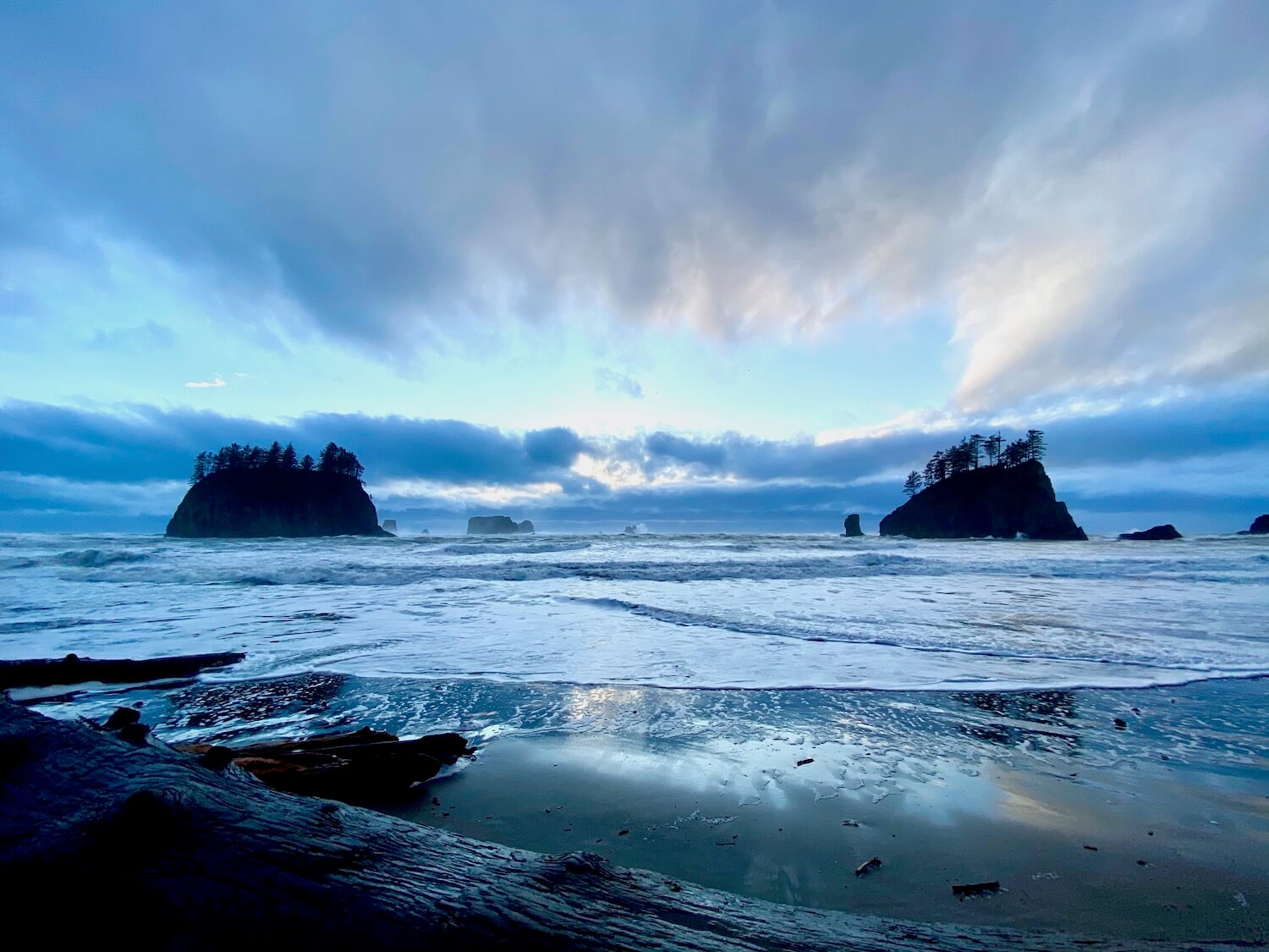 A beach scene of the Pacific Ocean waves crashing on the fine sandy beach with two large rocky islands in the background with fir trees growing on top.