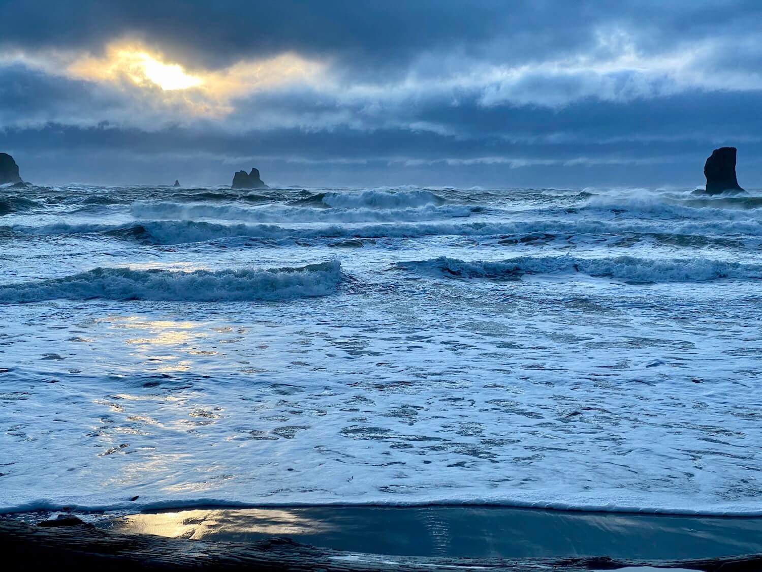  A Washington State beach scene with crashing waves on the sand of the coast. Several single rocks pop up in the background as the sun shines through dark storm clouds.
